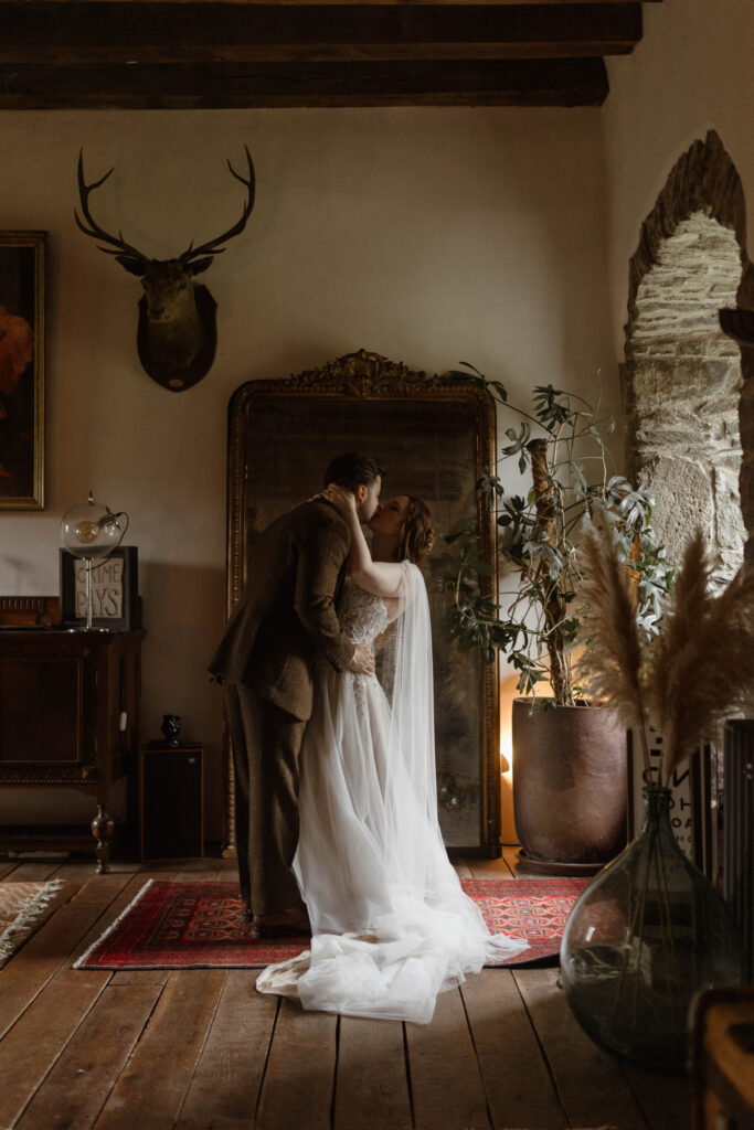 Romantic indoor portrait of Bethany and Adam kissing beside an antique mirror and stone archway inside Kilmartin Castle, captured in a fine-art, moody style.