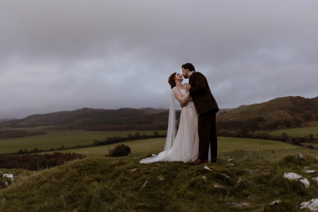 Bethany and Adam sharing a kiss on Dunadd Fort at dusk, with misty Argyll hills behind them, photographed in a cinematic, moody style during their Kilmartin Castle wedding.