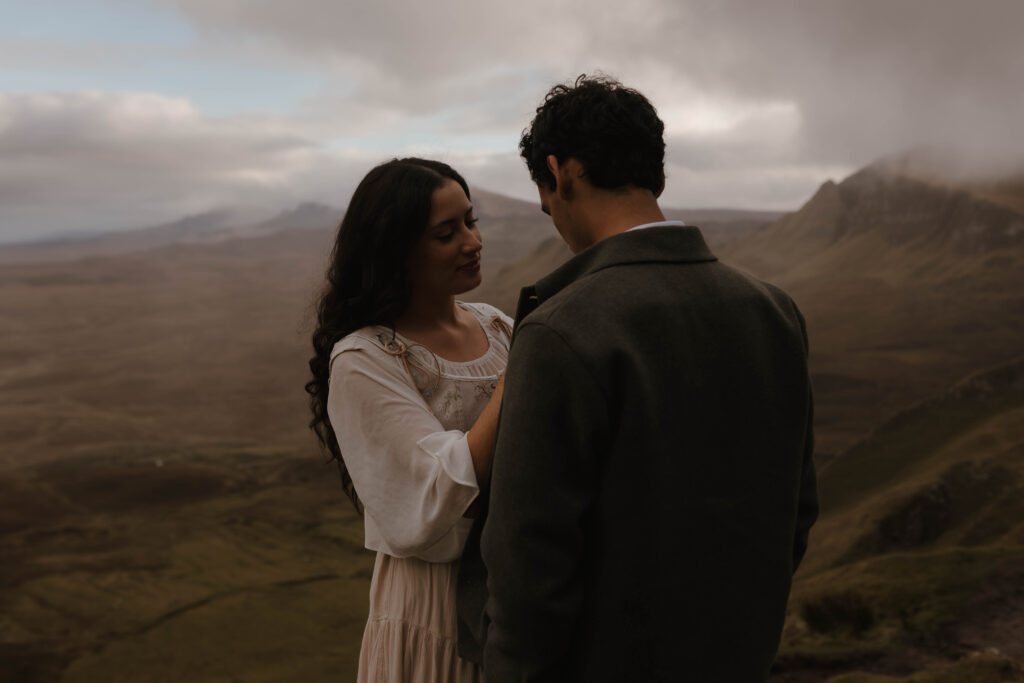Fine art portrait of a couple during an Isle of Skye photoshoot, with the woman adjusting her partner’s coat in soft, moody Highland light.