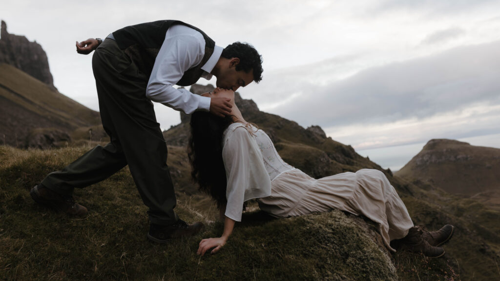 Cinematic couples portrait on the Isle of Skye, with a man leaning down to kiss his partner on a mossy hillside of the Quiraing, captured by Scotland fine art photographer Aly Robinson.