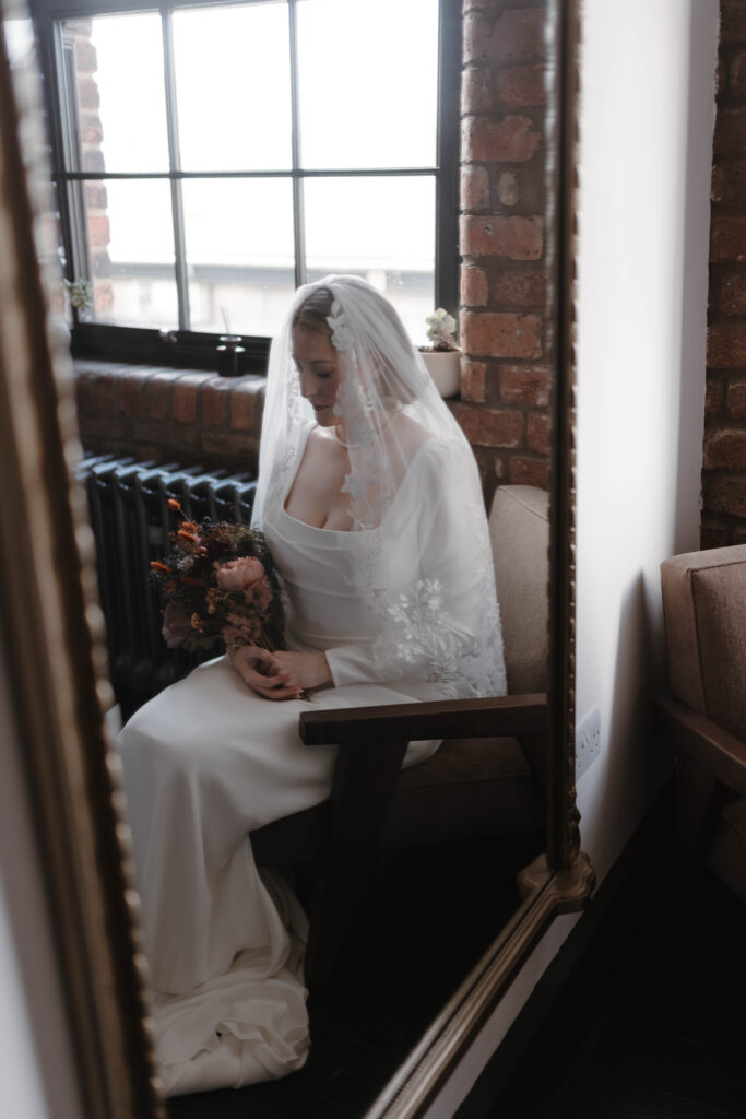 Reflective bridal portrait of a bride seated beside a tall mirror in soft morning light, photographed by fine art Scotland wedding photographer Aly Robinson.