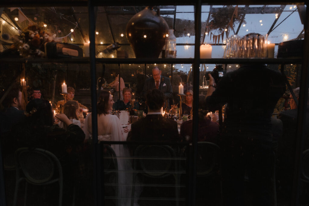 Atmospheric wedding dinner viewed through a glass greenhouse wall, glowing with candles and warm evening light, captured by fine art photographer Aly Robinson in Scotland.