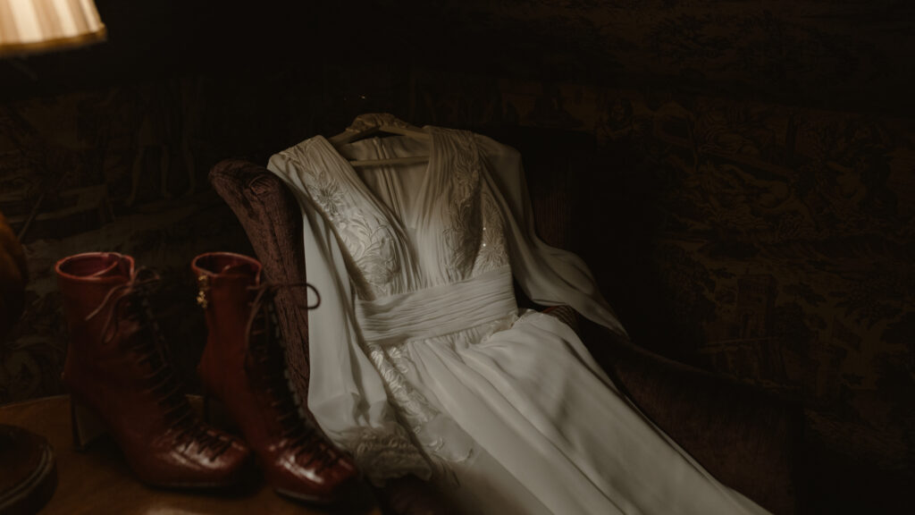 Embroidered silk wedding dress draped over vintage armchair with lace up leather boots inside Prestonfield House Edinburgh during luxury cinematic wedding photography session
