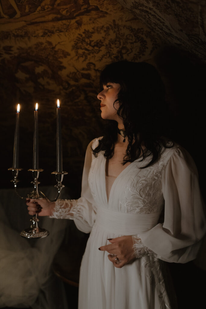 Bride in embroidered silk gown holding silver candelabra against historic tapestry walls at Prestonfield House Edinburgh for gothic inspired wedding photography