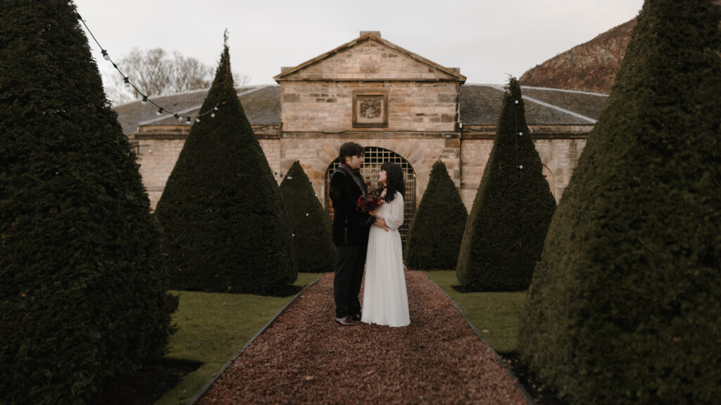 Bride and groom standing in the formal gardens at Prestonfield House Edinburgh with historic stone architecture for cinematic luxury wedding photography in Scotland