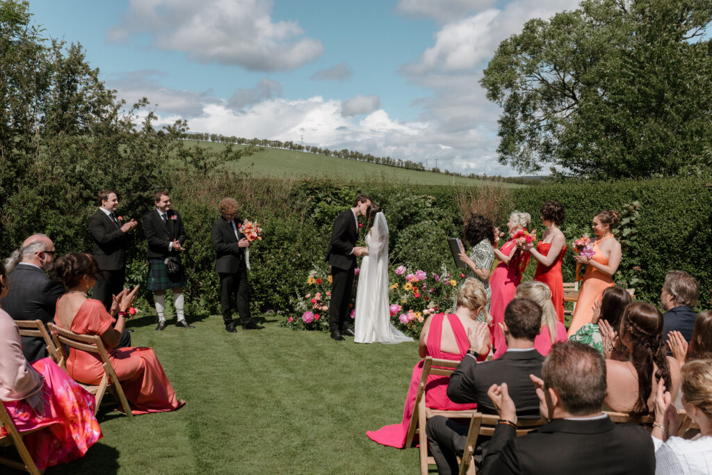 Outdoor summer wedding ceremony at Dalduff Farm near Glasgow with colourful guests, flowers and countryside views